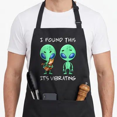 Vibrating cat apron - Two green aliens holding an orange tabby cat with white text above and below — shown on a black cotton canvas apron against a white background