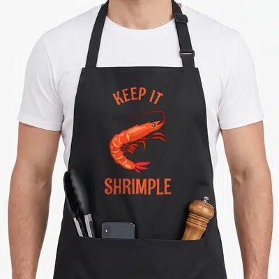 Shrimple shrimp keeping apron - Orange cartoon shrimp with arched KEEP IT text above and SHRIMPLE below — shown on a black cotton canvas apron against a white background