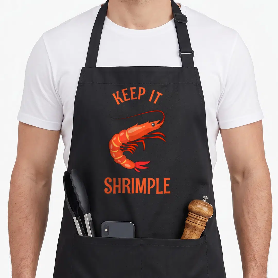 Shrimple shrimp keeping apron - Orange cartoon shrimp with arched KEEP IT text above and SHRIMPLE below — shown on a black cotton canvas apron against a white background