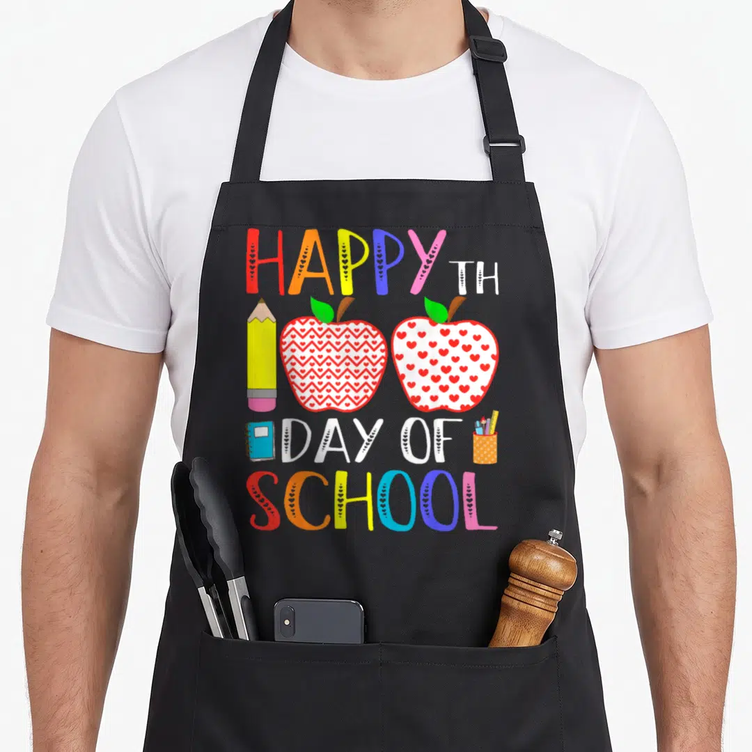 Funny 100th teacher apron - HAPPY TH 100 DAY OF SCHOOL text with cartoon pencil, two patterned apples, notebook — shown on a black cotton canvas apron against a white background