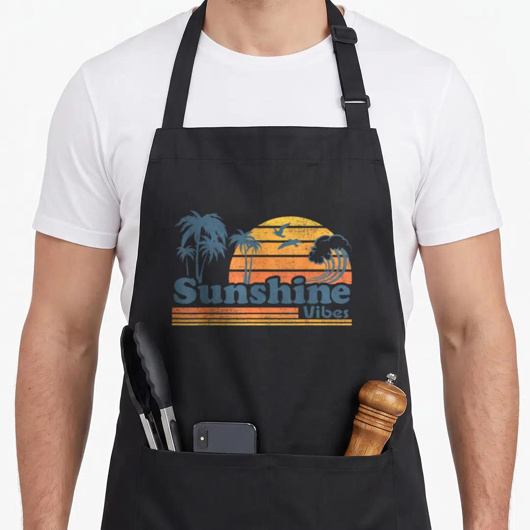 Sunshine 70s apron - Retro sun with stripes, palm trees, birds, wave, and blue-grey text reading Sunshine Vibes — shown on a black cotton canvas apron against a white background