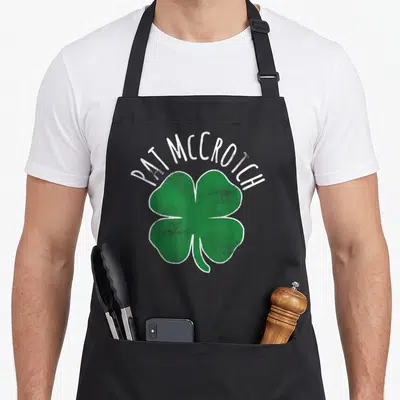 Pat the dirty apron - PAT MCCROTCH text above green four-leaf clover with distressed white outline — shown on a black cotton canvas apron against a white background