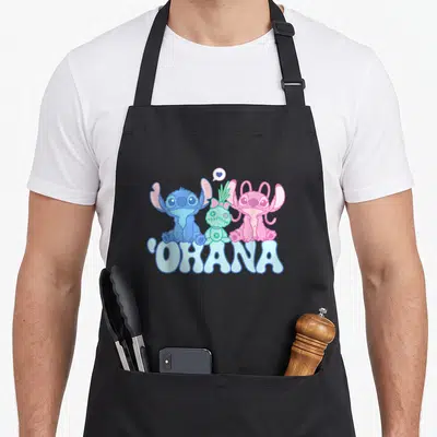 Scrump angels in aprons - Three cartoon aliens in blue pink green sit above OHANA text with heart bubble — shown on a black cotton canvas apron against a white background