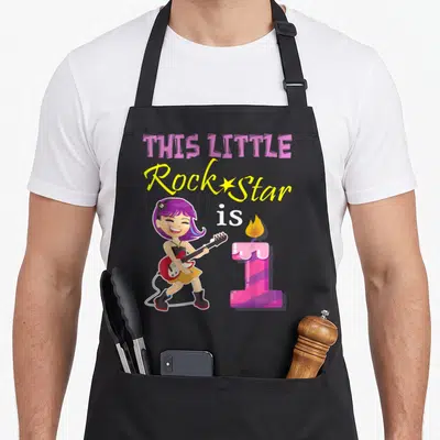 Rock first birthday apron - Cartoon girl with purple hair plays guitar next to pink number 1 candle under stacked text — shown on a black cotton canvas apron against a white background