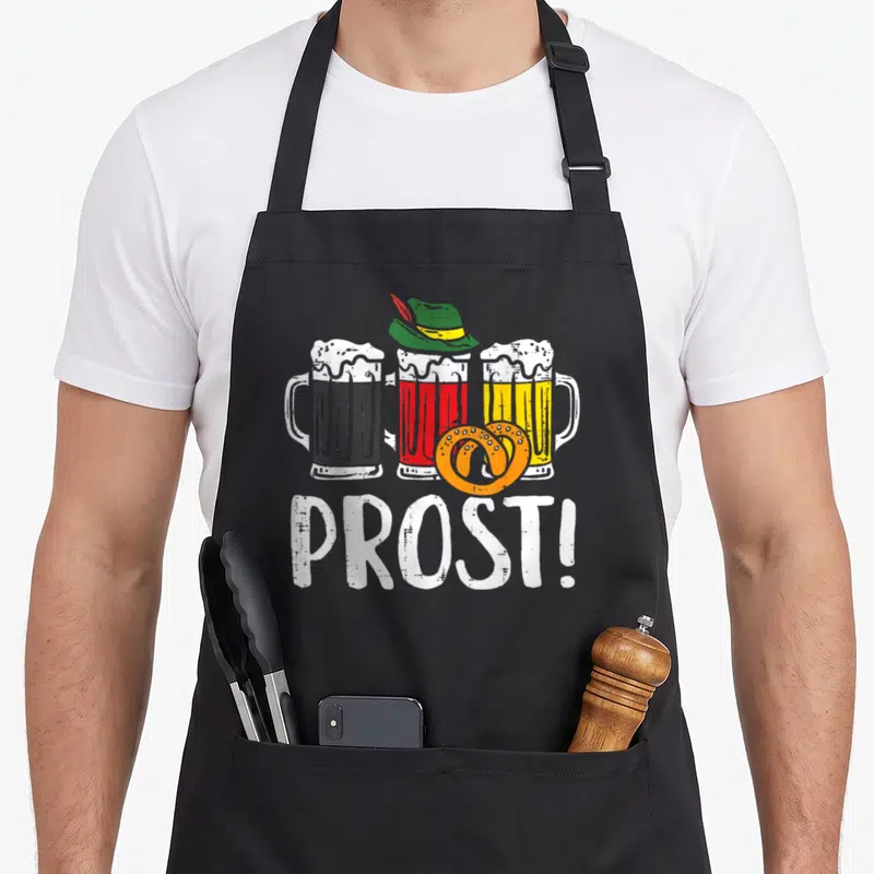 Prost german oktoberfest aprons - Three beer mugs with hat and orange pretzel above white PROST! text — shown on a black cotton canvas apron against a white background
