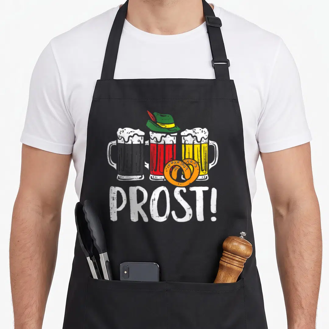 Prost german oktoberfest aprons - Three beer mugs with hat and orange pretzel above white PROST! text — shown on a black cotton canvas apron against a white background