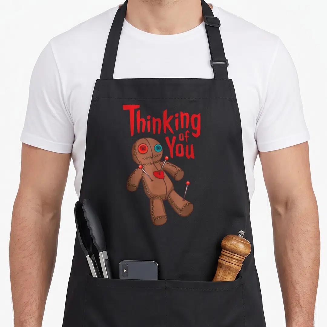 Wiccan doll apron - Red 'Thinking of You' text above brown voodoo doll with pins and mismatched button eyes — shown on a black cotton canvas apron against a white background