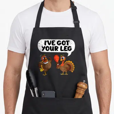 Leg fall aprons - Two cartoon turkeys under speech bubble text holding a cooked drumstick — shown on a black cotton canvas apron against a white background