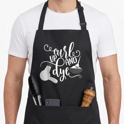 Curl hair dye apron - White cursive text curl UP AND dye with hair dryer and dye bowl on black — shown on a black cotton canvas apron against a white background