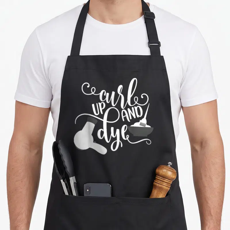 Curl hair dye apron - White cursive text curl UP AND dye with hair dryer and dye bowl on black — shown on a black cotton canvas apron against a white background