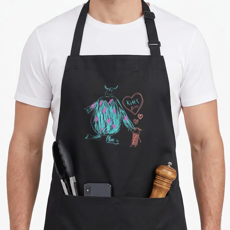 Sulley kitty aprons - Blue monster holding pink child hand with heart reading KITTY BOO in line art — shown on a black cotton canvas apron against a white background