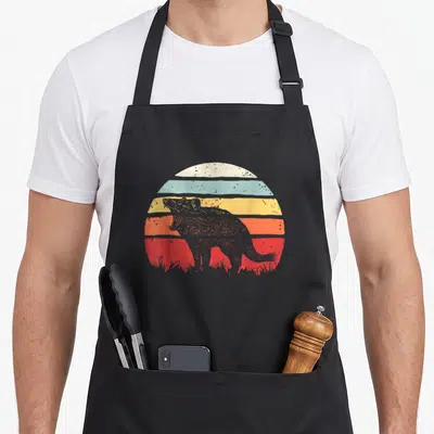 Catch devil's apron - Grey Tasmanian devil silhouette over red orange yellow blue cream striped sun with grass — shown on a black cotton canvas apron against a white background