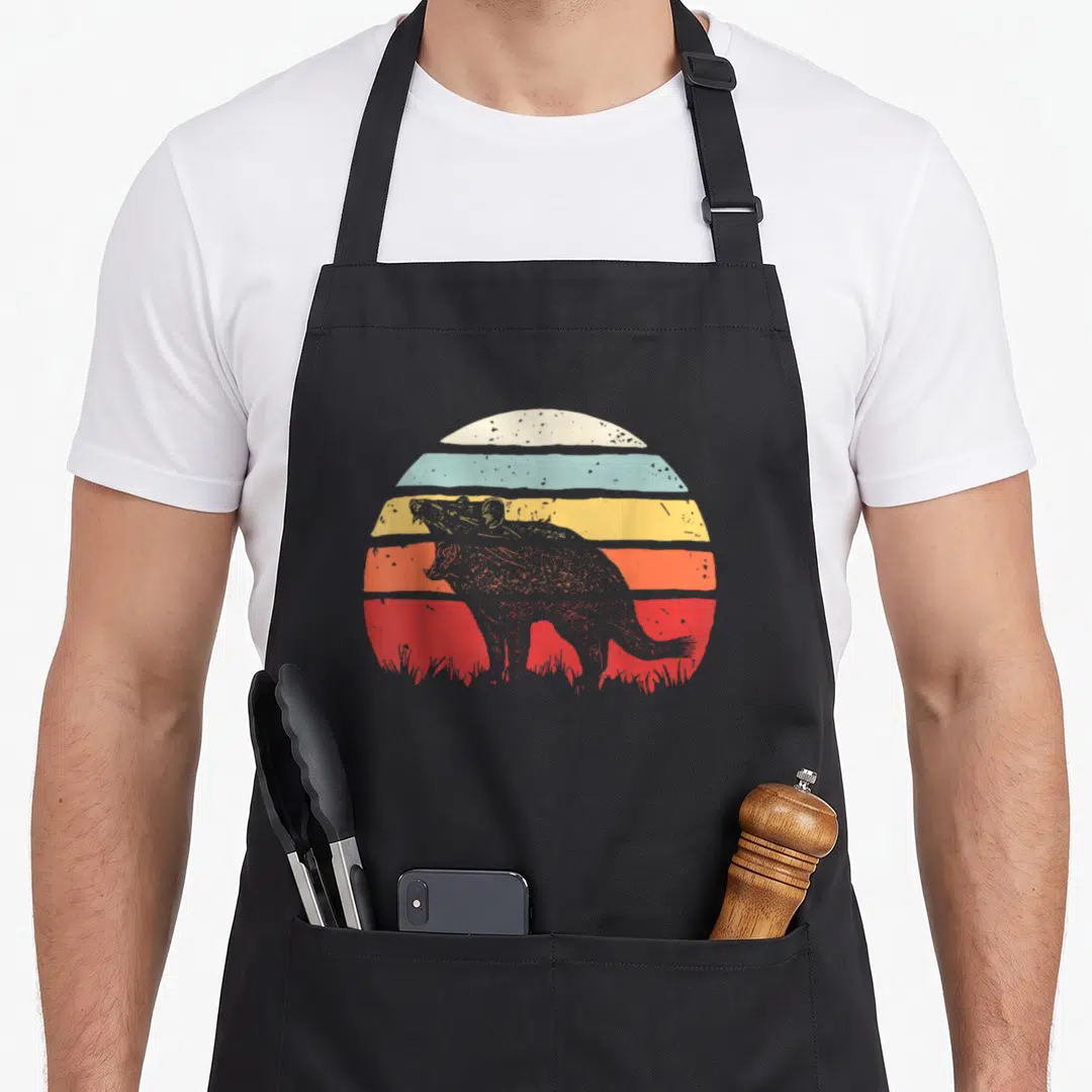 Catch devil's apron - Grey Tasmanian devil silhouette over red orange yellow blue cream striped sun with grass — shown on a black cotton canvas apron against a white background