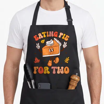 Eating thanksgiving apron - Cartoon orange pie slice with EATING PIE FOR TWO text and autumn leaves in red yellow brown — shown on a black cotton canvas apron against a white background