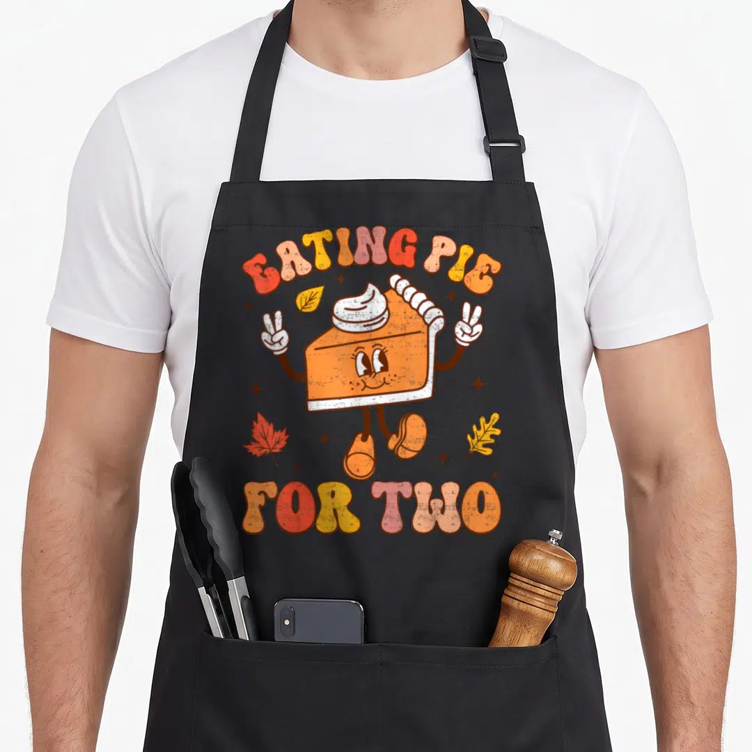Eating thanksgiving apron - Cartoon orange pie slice with EATING PIE FOR TWO text and autumn leaves in red yellow brown — shown on a black cotton canvas apron against a white background