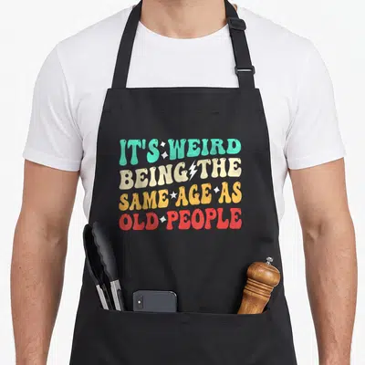 Cool sarcastic weird aprons - Stacked retro text in teal beige orange and red with white stars and lightning bolt — shown on a black cotton canvas apron against a white background