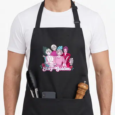 Palm golden apron - Stay Golden text below four cartoon women with pink roses and blue leaves — shown on a black cotton canvas apron against a white background
