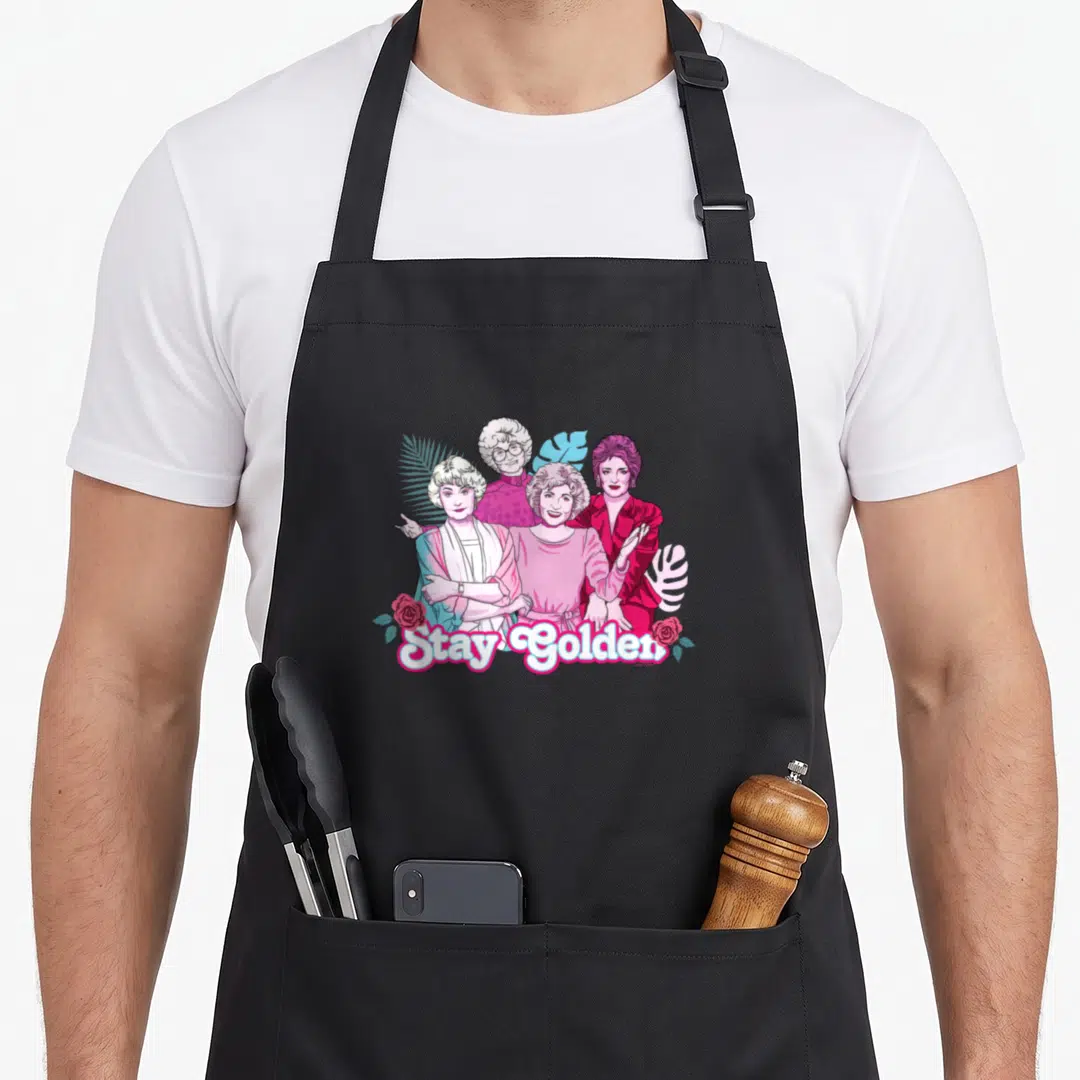 Palm golden apron - Stay Golden text below four cartoon women with pink roses and blue leaves — shown on a black cotton canvas apron against a white background