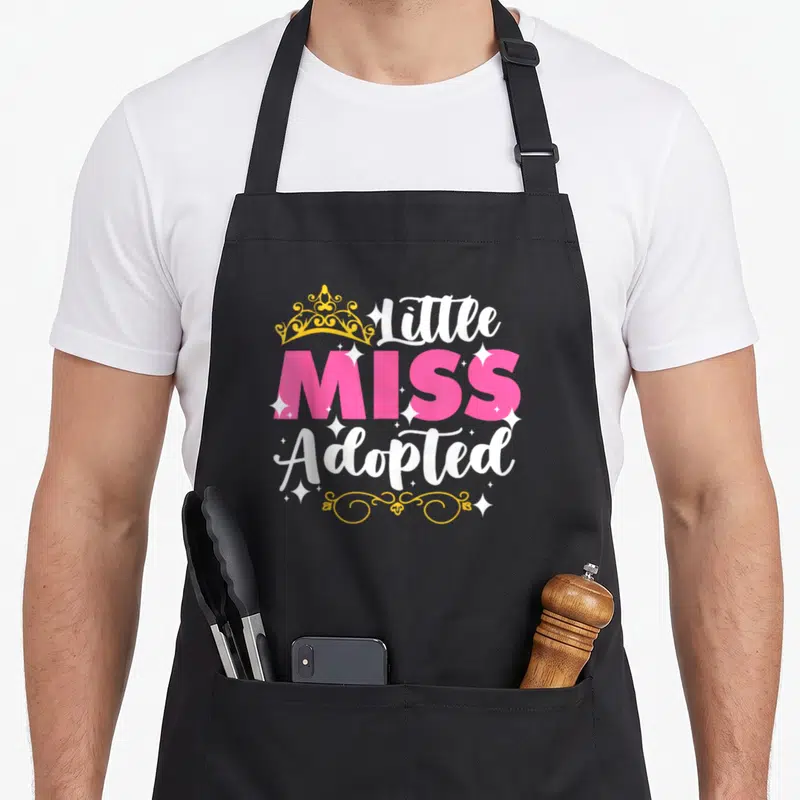 Miss days christmas apron - Little Miss Adopted text with yellow crown and white sparkles on pink background — shown on a black cotton canvas apron against a white background