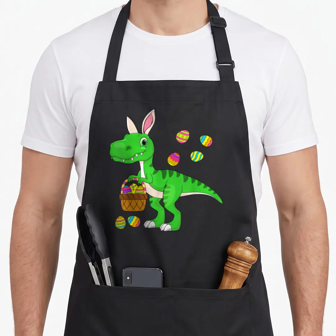 Dinosaur basket apron - Green cartoon dinosaur with pink bunny ears holding a basket of colorful eggs — shown on a black cotton canvas apron against a white background