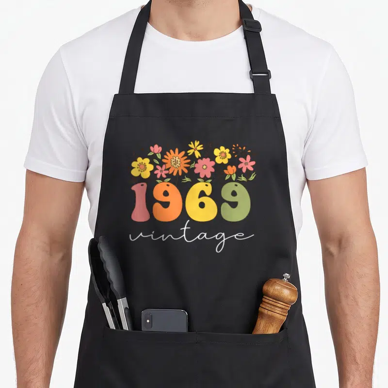 54th floral aprons - Retro 1969 vintage text with yellow orange pink green and white flowers growing from numbers — shown on a black cotton canvas apron against a white background