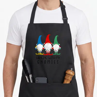 Gnomy christmas gnome apron - Three cartoon gnomes in blue red and green hats above hanging with my gnomies text — shown on a black cotton canvas apron against a white background