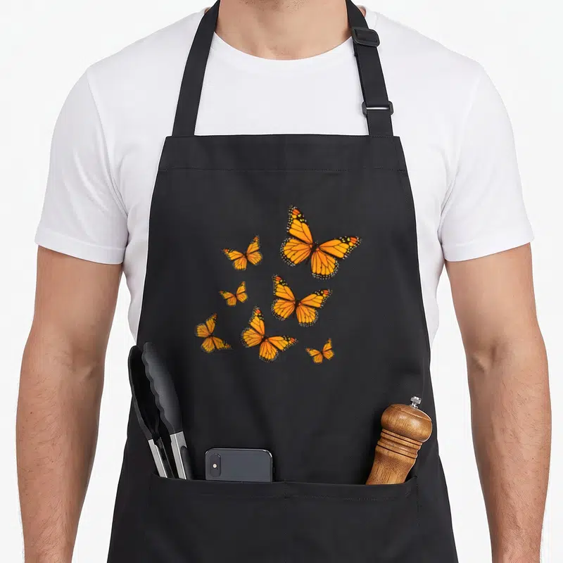 Cute monarch butterfly apron - Six orange and black monarch butterflies with white dots scattered across grey background — shown on a black cotton canvas apron against a white background