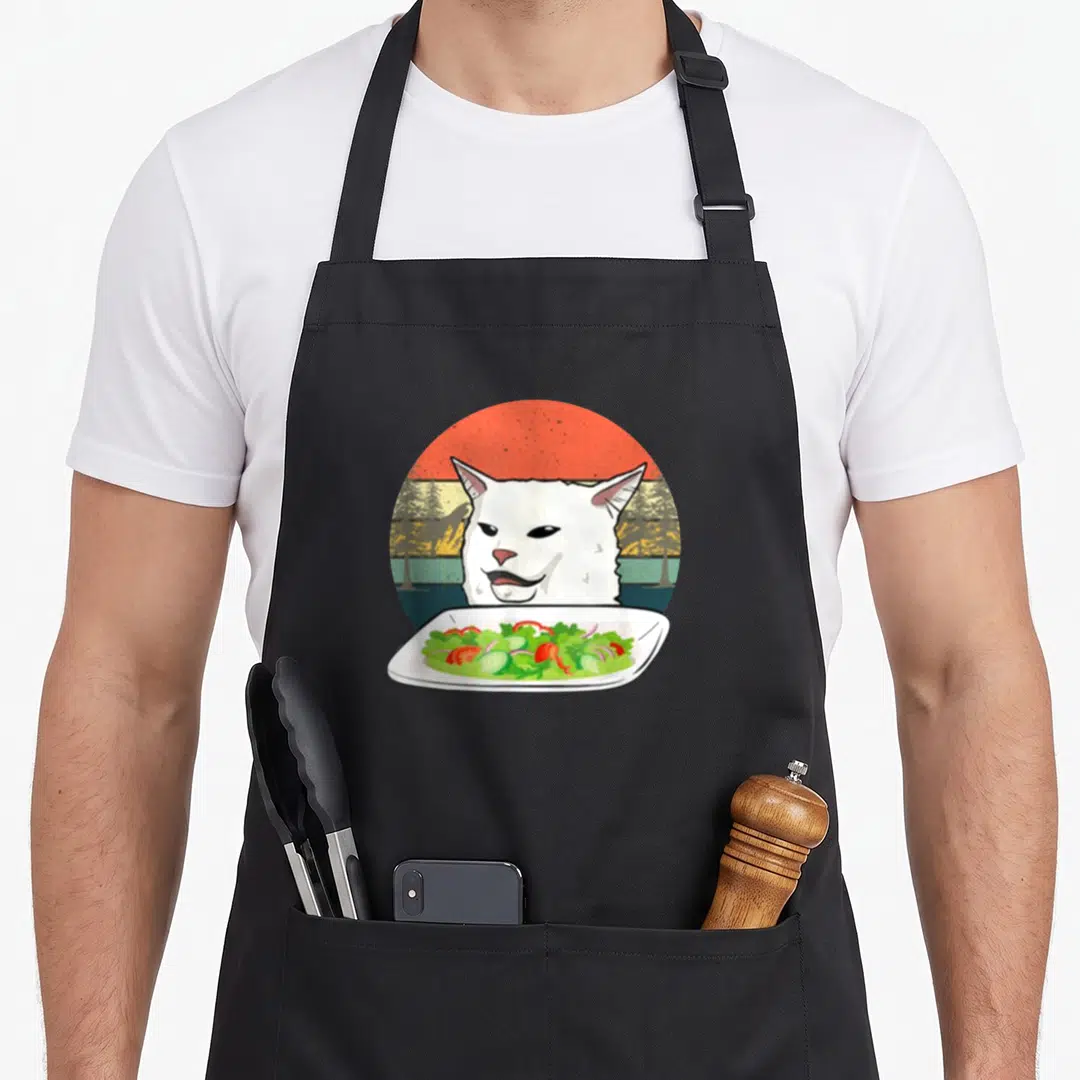 Cat table apron - Cartoon white cat face behind plate of salad with orange sun and green trees — shown on a black cotton canvas apron against a white background