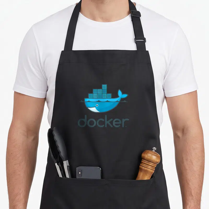 Docker devops apron - Blue cartoon whale carrying stacked shipping containers above dark grey docker text — shown on a black cotton canvas apron against a white background