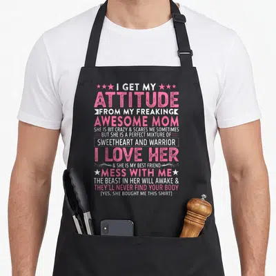 Freaking attitude aprons - Stacked pink and white text reading I get my attitude from my freaking awesome mom — shown on a black cotton canvas apron against a white background