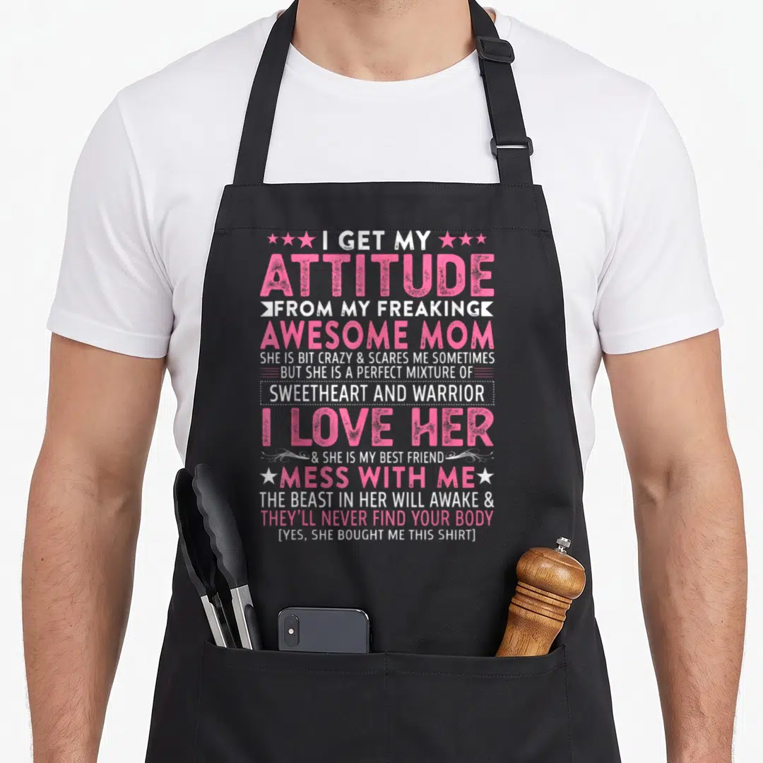 Freaking attitude aprons - Stacked pink and white text reading I get my attitude from my freaking awesome mom — shown on a black cotton canvas apron against a white background
