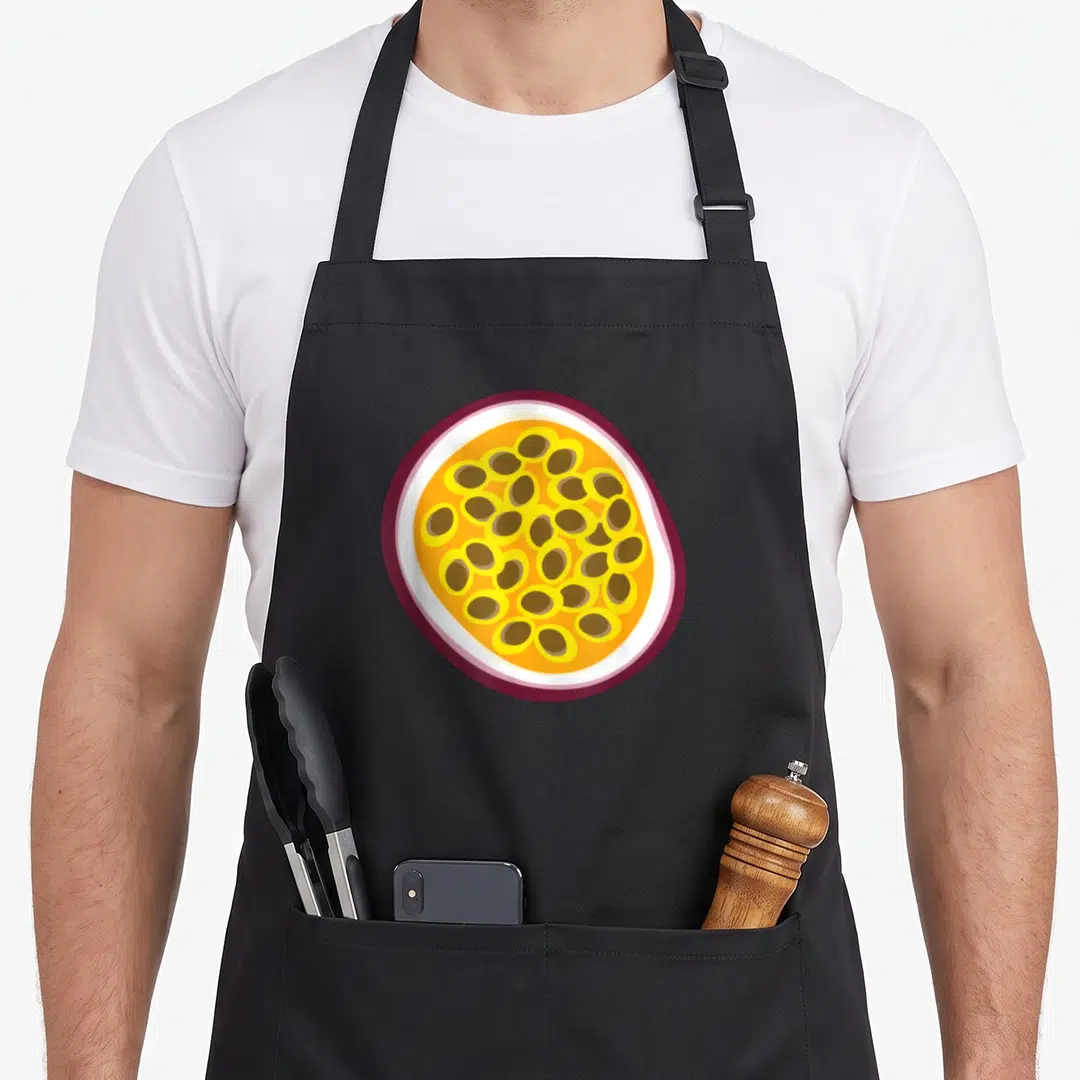 Passion fruit apron - Cartoon passion fruit cross-section showing purple rind, white pith, yellow pulp, and brown seeds — shown on a black cotton canvas apron against a white background
