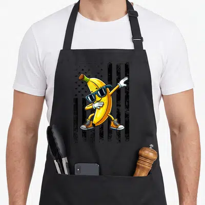 Dabbing banana apron - Cartoon yellow banana wearing sunglasses and orange shoes posing in front of a black and grey — shown on a black cotton canvas apron against a white background