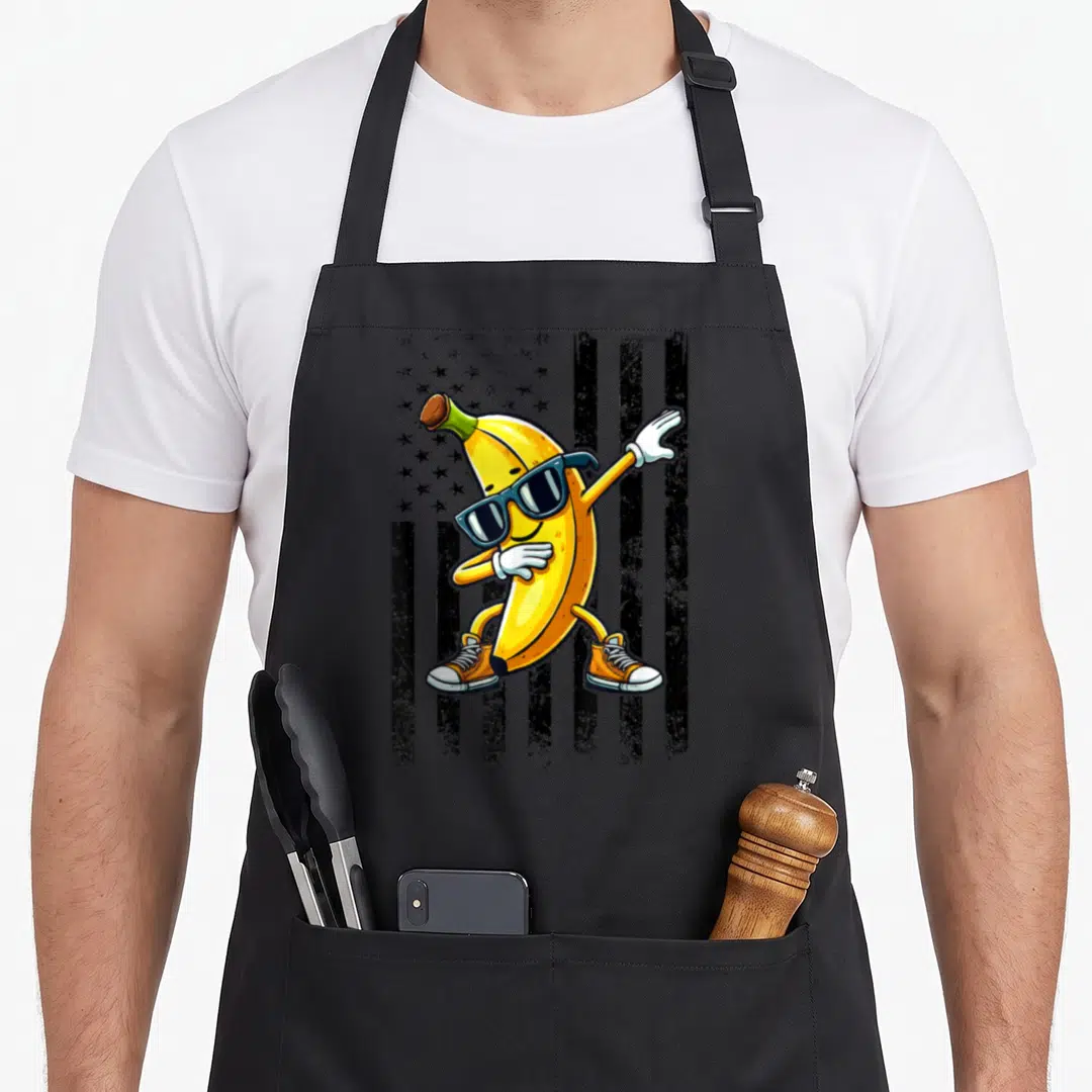 Dabbing banana apron - Cartoon yellow banana wearing sunglasses and orange shoes posing in front of a black and grey — shown on a black cotton canvas apron against a white background