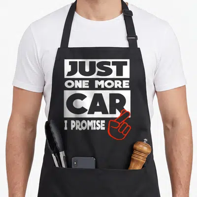 Nur auto apron - Stacked text reading Just One More Car I Promise with red outlined crossed fingers on grey background — shown on a black cotton canvas apron against a white background