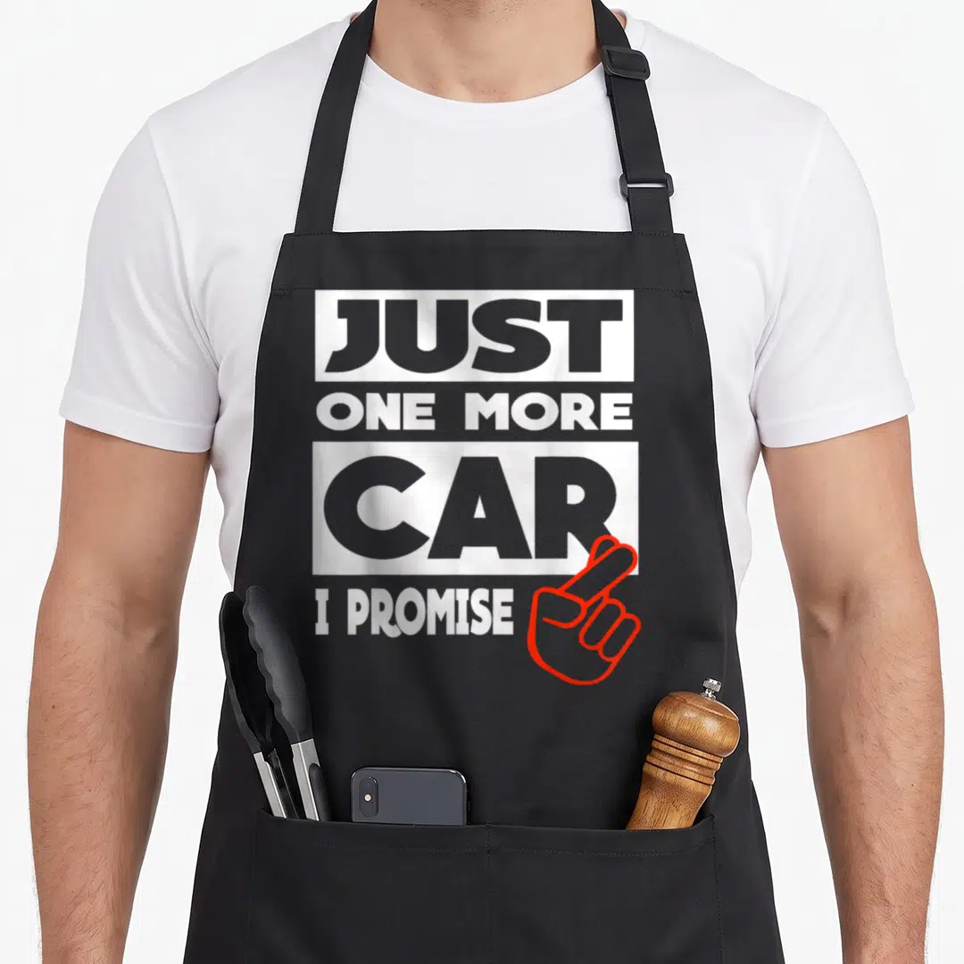 Nur auto apron - Stacked text reading Just One More Car I Promise with red outlined crossed fingers on grey background — shown on a black cotton canvas apron against a white background