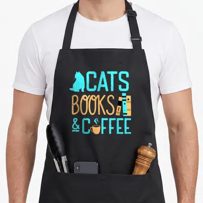 Nerd cat apron - Cyan cat silhouette and tan coffee cup flanking stacked text reading CATS BOOKS & COFFEE — shown on a black cotton canvas apron against a white background