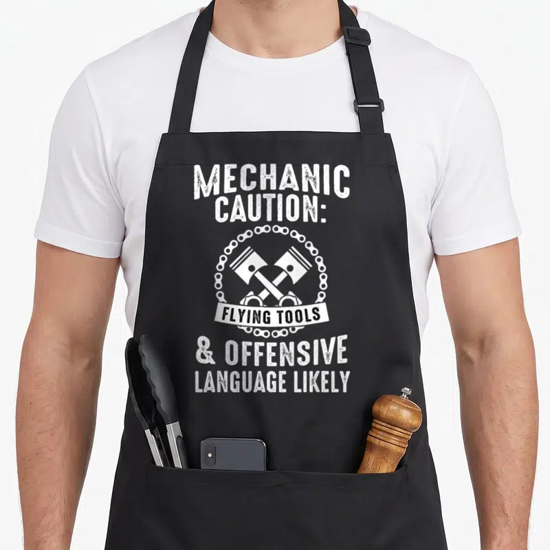 Cute car auto mechanic apron - MECHANIC CAUTION text above two crossed hammers in chain with FLYING TOOLS banner below — shown on a black cotton canvas apron against a white background