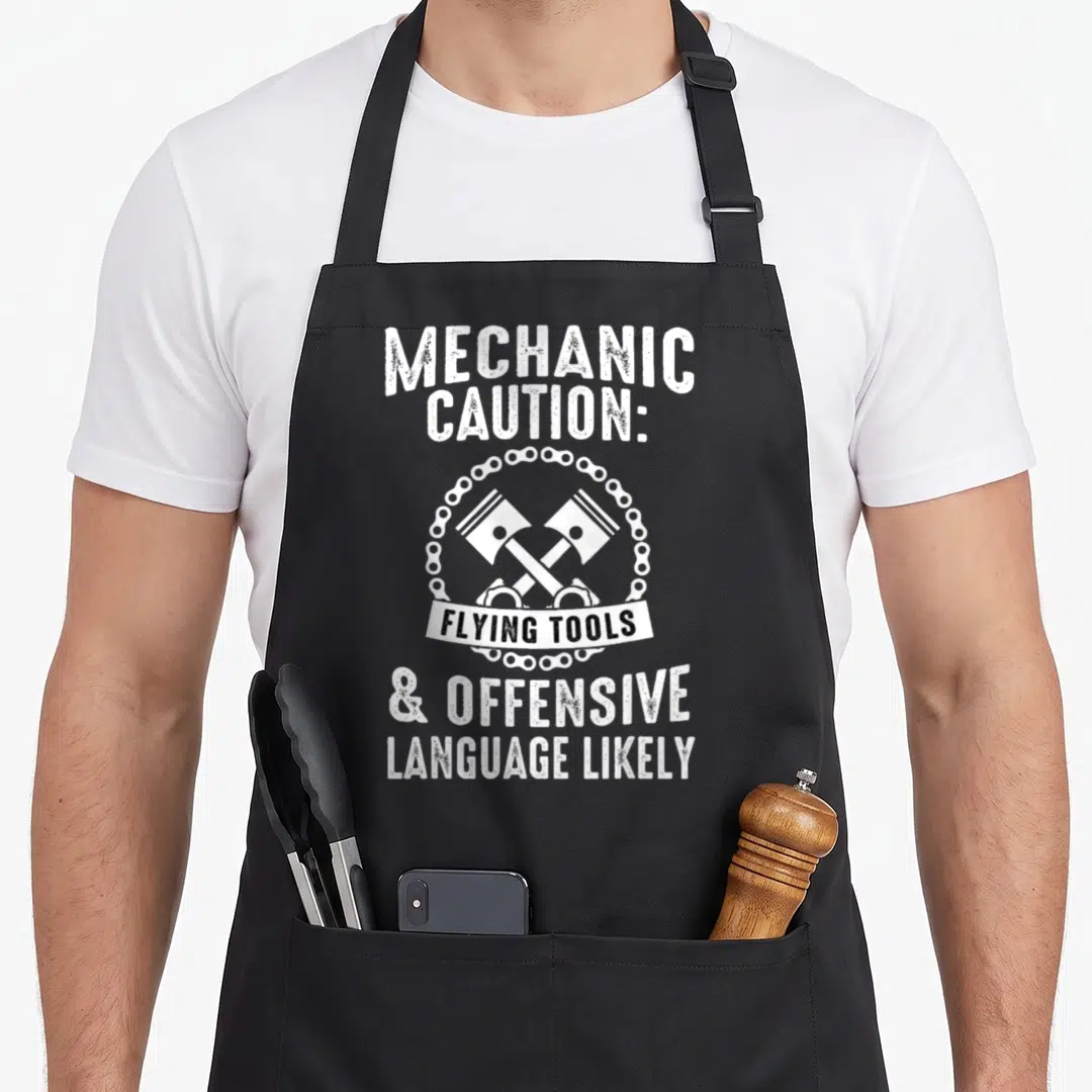 Cute car auto mechanic apron - MECHANIC CAUTION text above two crossed hammers in chain with FLYING TOOLS banner below — shown on a black cotton canvas apron against a white background