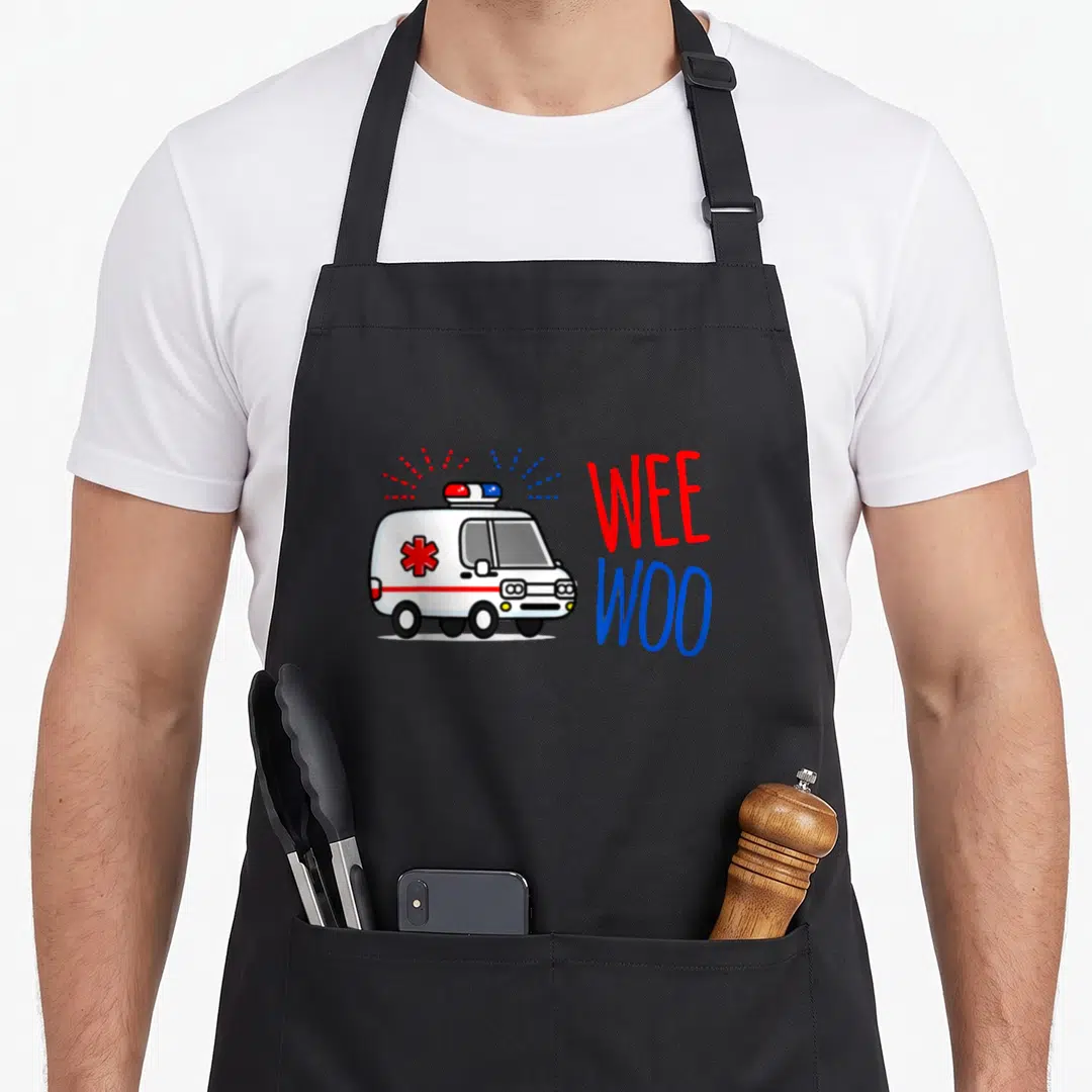 Wee future paramedic apron - Cartoon ambulance with red blue sound lines and stacked Wee Woo text — shown on a black cotton canvas apron against a white background