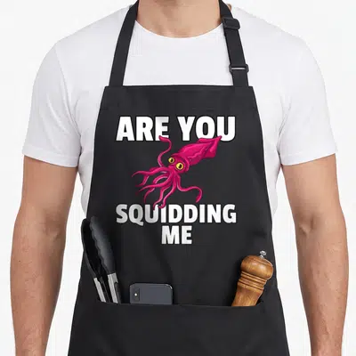 Squidding squid mom apron - Pink cartoon squid with yellow eyes framed by white Are You Squidding Me text — shown on a black cotton canvas apron against a white background