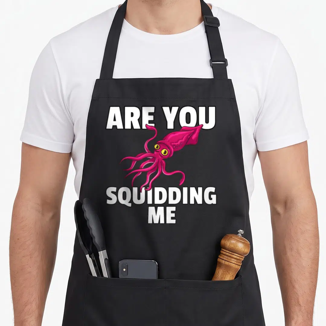 Squidding squid mom apron - Pink cartoon squid with yellow eyes framed by white Are You Squidding Me text — shown on a black cotton canvas apron against a white background