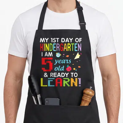 1st learning apron - Stacked text My 1st Day of Kindergarten with cartoon clock apple and paper airplane icons — shown on a black cotton canvas apron against a white background