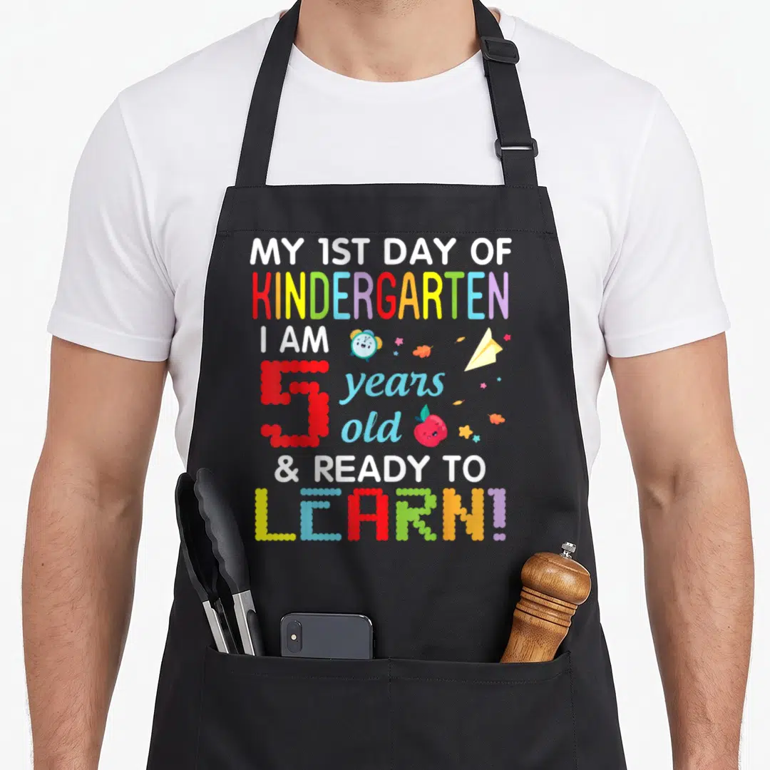 1st learning apron - Stacked text My 1st Day of Kindergarten with cartoon clock apple and paper airplane icons — shown on a black cotton canvas apron against a white background