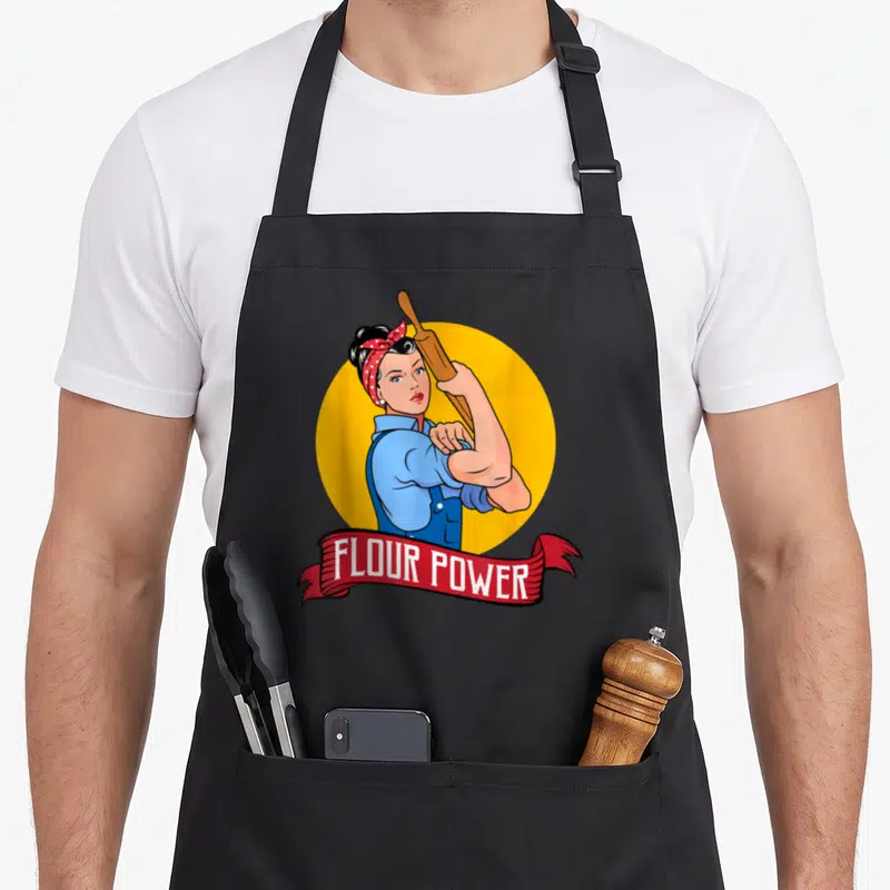 Flour baker apron - Retro woman flexing arm holding rolling pin with FLOUR POWER text on red ribbon — shown on a black cotton canvas apron against a white background