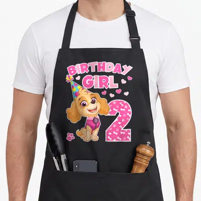 Skye birthday apron - Cartoon dog wearing party hat next to pink number 2 and BIRTHDAY GIRL text with hearts — shown on a black cotton canvas apron against a white background