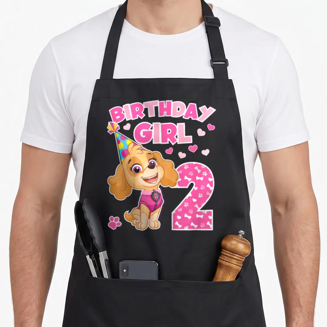 Skye birthday apron - Cartoon dog wearing party hat next to pink number 2 and BIRTHDAY GIRL text with hearts — shown on a black cotton canvas apron against a white background