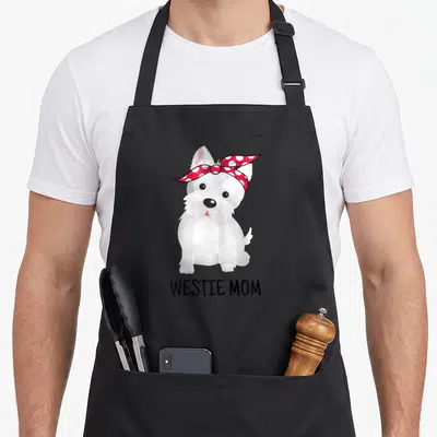 Westie dog apron - Cartoon white terrier with red bandana and black WESTIE MOM text — shown on a black cotton canvas apron against a white background