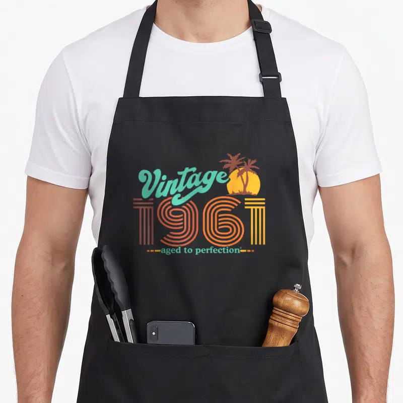 Aged perfection 60th birthday apron - Retro 1961 text design with teal script, orange numbers, and yellow sun palm trees — shown on a black cotton canvas apron against a white background