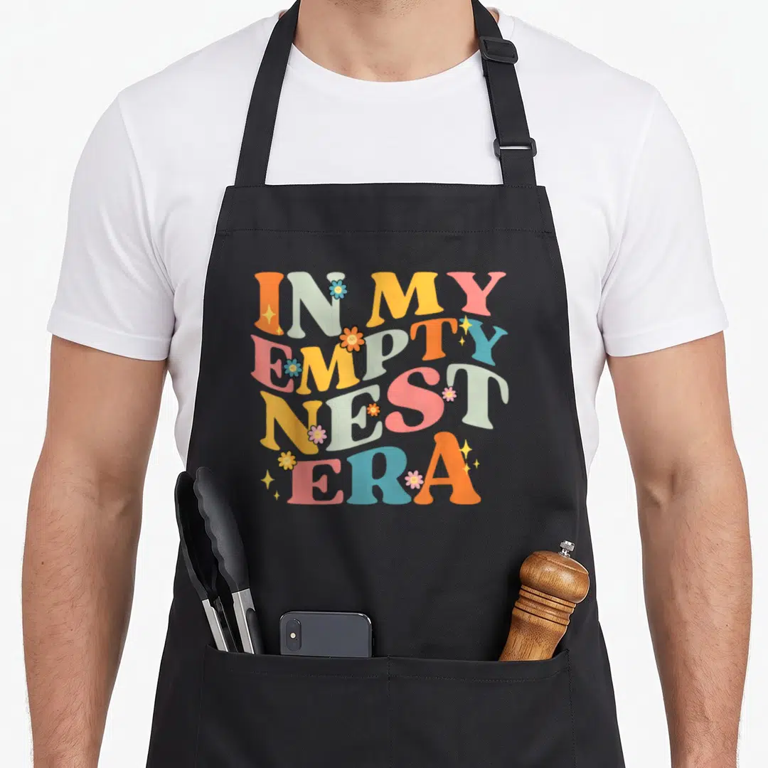 Empty mom era apron - Stacked IN MY EMPTY NEST ERA text with smiling flowers and sparkles in orange, grey, yellow — shown on a black cotton canvas apron against a white background